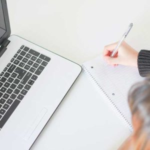 hand holding pen over notepad in front of laptop