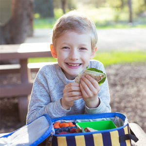 smiling young boy sitting in front of an open lunchbox and eating a sandwich