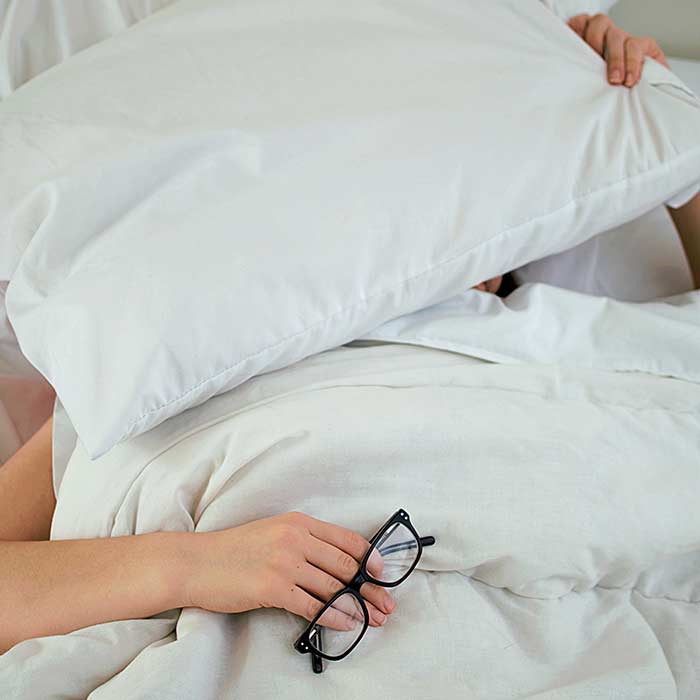 person in bed under white covers pulling pillow over head while other hand holds spectacles